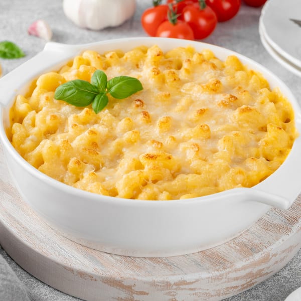 Baked mac and cheese in a white dish, garnished with basil, with tomatoes and garlic in the background.