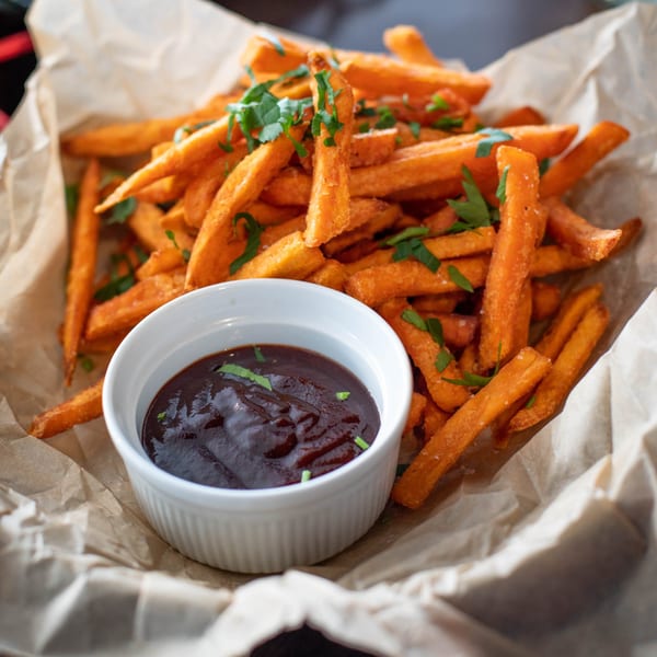 Sweet potato fries with green herbs and a side of dark dipping sauce in a white bowl.