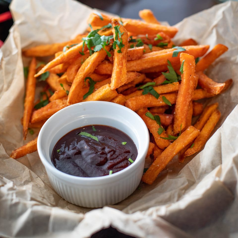 Sweet potato fries with green herbs and a side of dark dipping sauce in a white bowl.