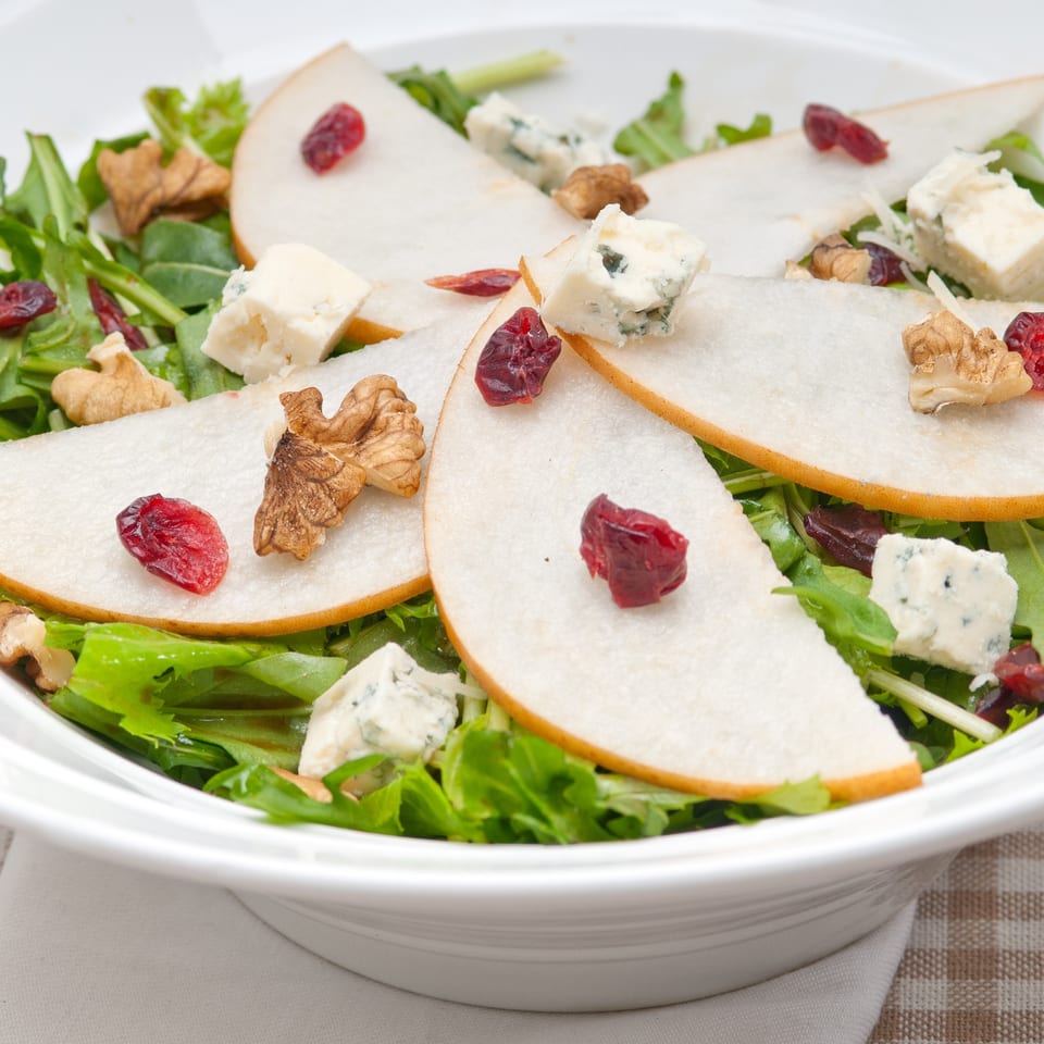 A fresh salad with pear slices, blue cheese, walnuts, and cranberries in a white bowl.