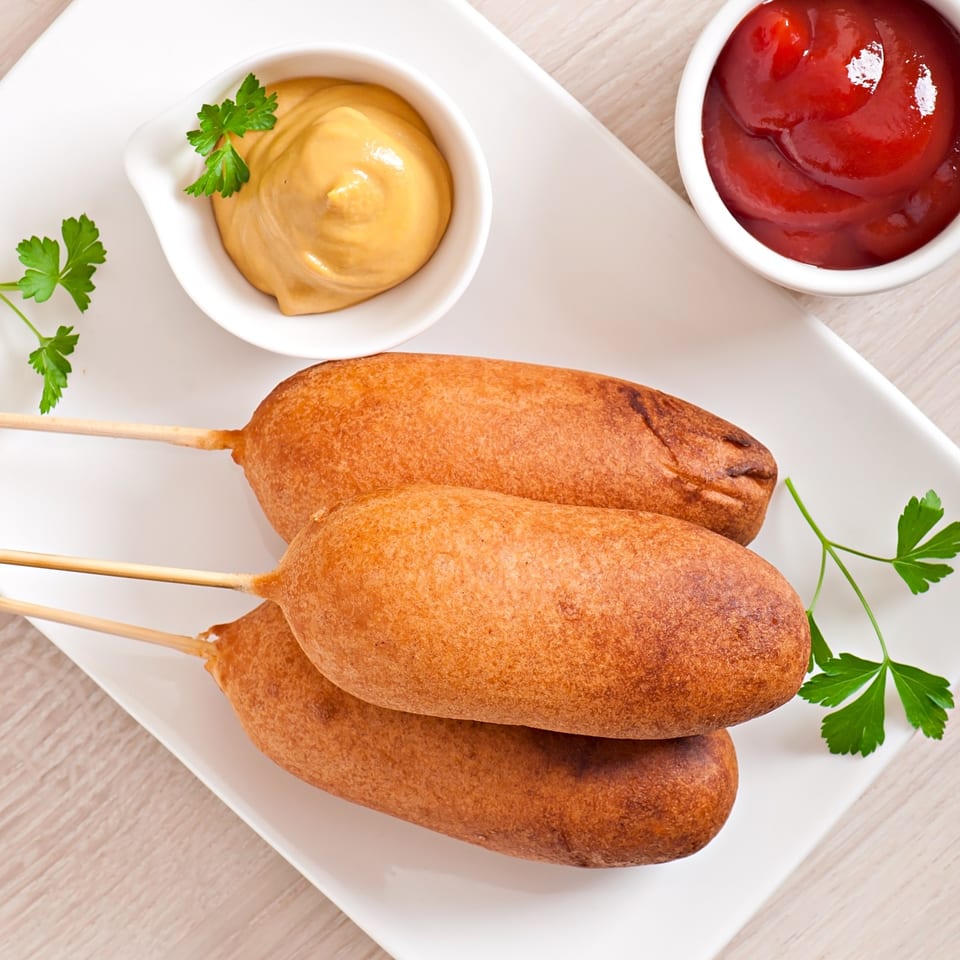 Three golden-brown corn dogs on a white plate, with bowls of mustard and ketchup, garnished with parsley.